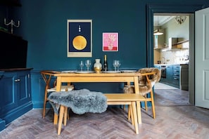 Un comedor con una mesa de madera dispuesta para cuatro personas, flanqueada por sillas con cojines de piel. Un cuadro enmarcado cuelga sobre la mesa, y se aprecia una cocina al fondo.