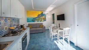 Een moderne keuken met een gasfornuis, een eethoek met tafel en stoelen, en een woonkamer met een bank en een schilderij aan de muur.