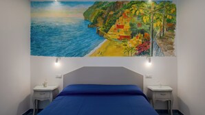 Een slaapkamer met een groot muurschildering van een kustdorp, een bed met een blauwe spreidek en twee nachtkastjes met lampen.