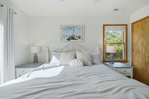 Un dormitorio ordenado con cama, dos mesitas de noche, una mesita auxiliar, una ventana con vistas y una puerta.