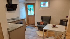 Ett kompakt kök med en inbyggd spis, en matplats med bord och stolar, en grå fåtölj och ett litet sidobord.