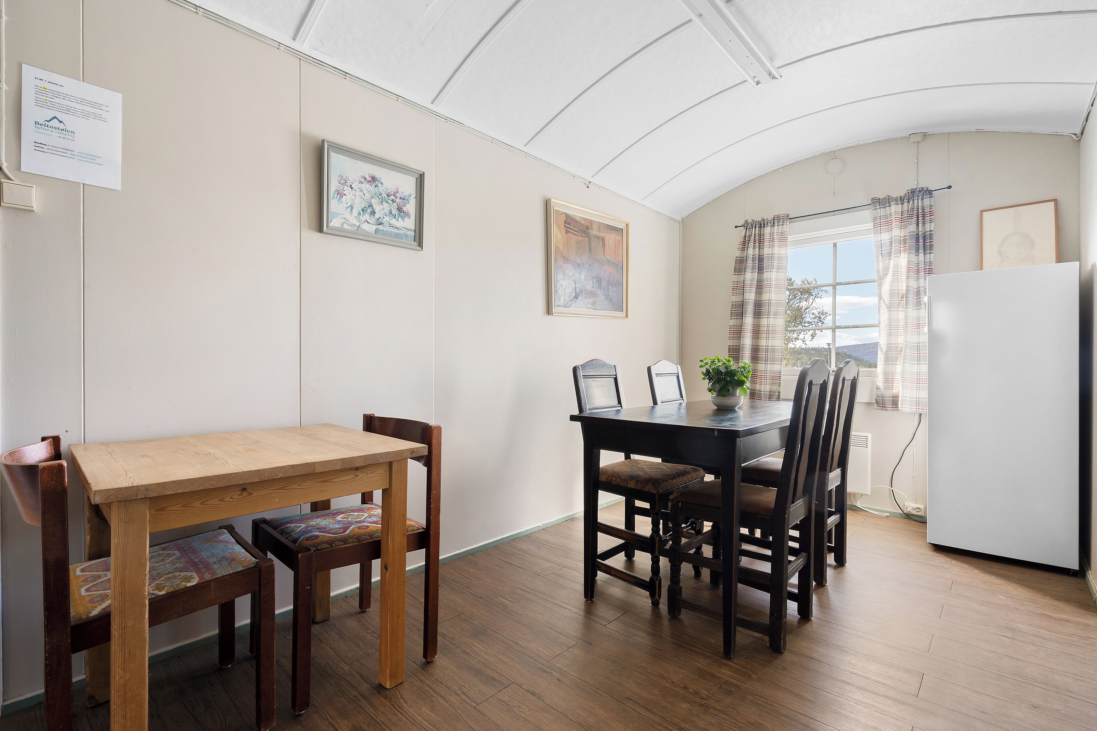 A dining area with a wooden table and chairs, a white refrigerator, and a window with curtains.