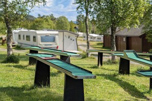 Mesas al aire libre con tapas de fieltro verde, instaladas en un área cubierta de césped con árboles y vehículos recreativos al fondo.