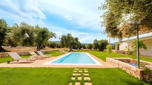 Una piscina rectangular con borde de piedra, rodeada por un césped cuidado y algunos árboles.