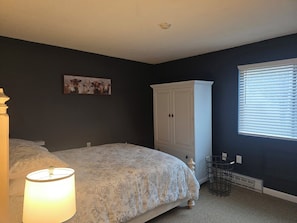 A bedroom with a bed, a nightstand, a dresser, a window with blinds, and a ceiling light.