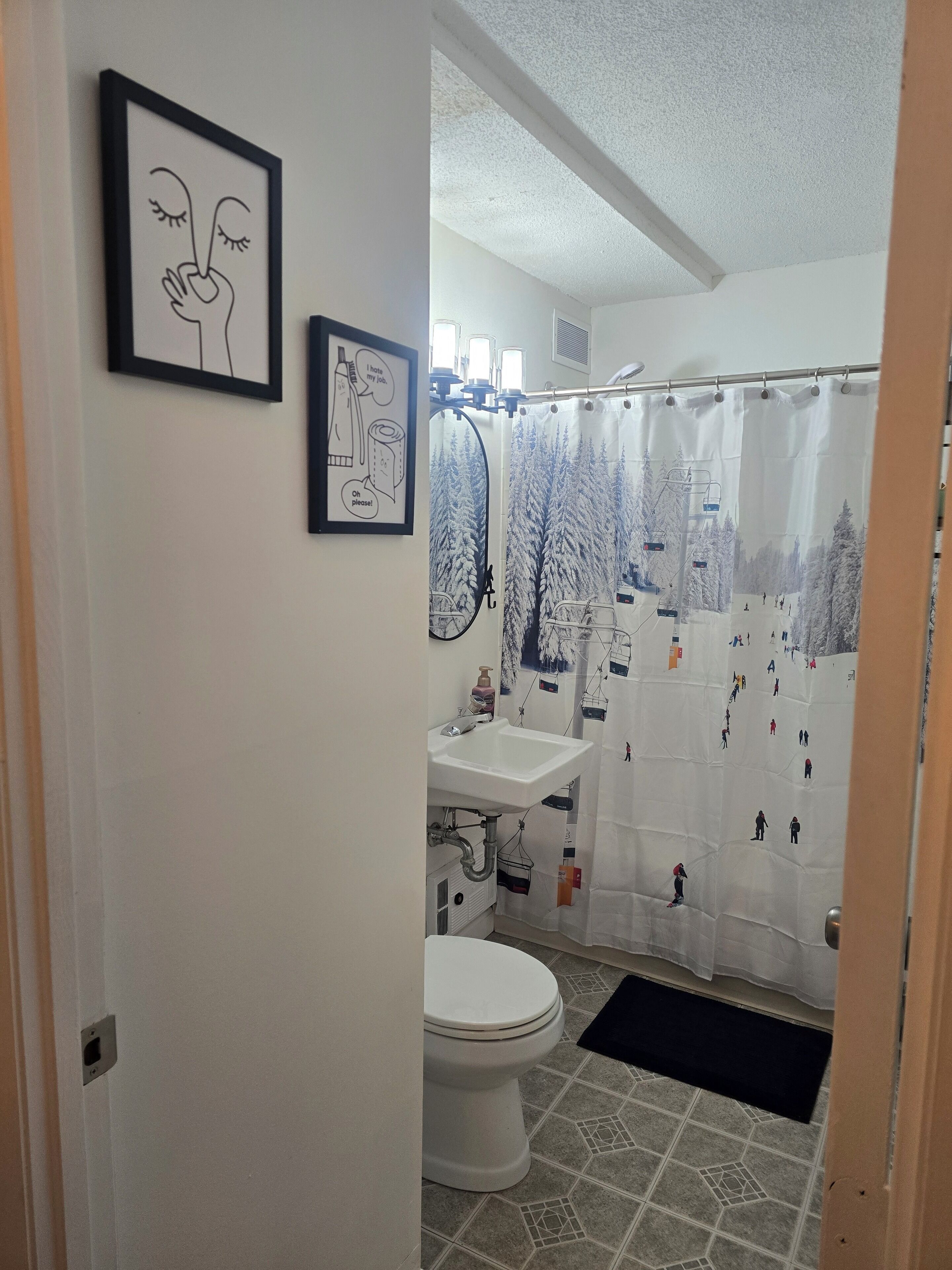 A bathroom with a white toilet, a sink, and a shower curtain depicting a snowy mountain scene. There are two framed pictures on the wall.
