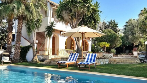 A swimming pool with lounge chairs and an umbrella, surrounded by palm trees and a building with arched windows. - VFH The Sicilian Dream Pool Villa (Altavilla Milicia)
