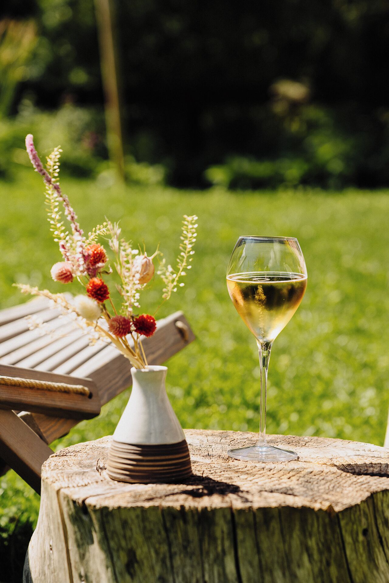 Una copa de vino blanco sobre un tocón de madera, junto a un florero con flores.