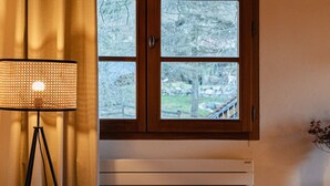 Een gezellige kamer met een stoel, een klein tafeltje met een vaas, een vloerlamp en een raam met uitzicht op bomen.