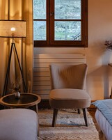 Una habitación acogedora con una silla, una mesita con un florero, una lámpara de pie y un ventanal con vistas a árboles.