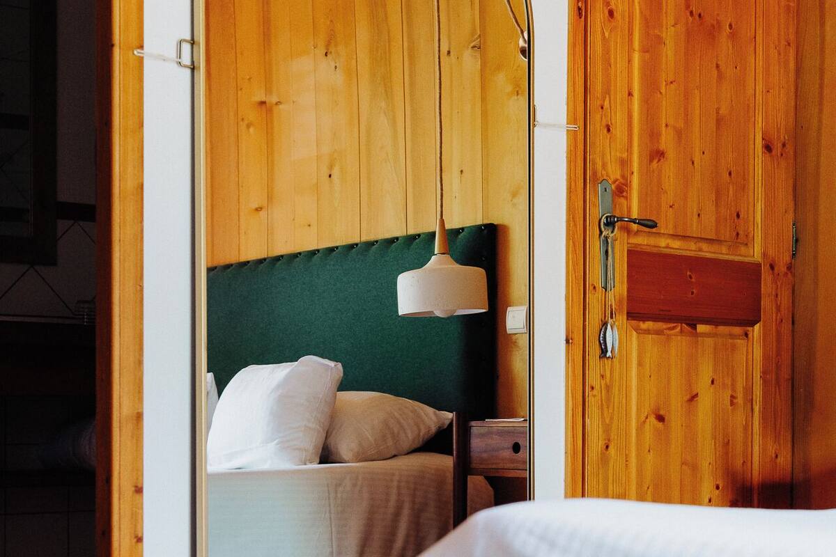 Une chambre avec une porte en bois, un lit avec une literie blanche, une tête de lit recouverte d’un tissu vert et un luminaire suspendu.