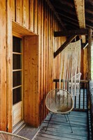 Una cabaña de madera con un ventanal grande, un sillón de macramé y una silla tejida en una terraza de madera.