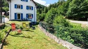 Una casa de dos pisos con fachada blanca y contraventanas azules, patio cercado con sillas y montañas al fondo.