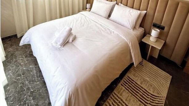 Un lit bien fait, avec du linge de lit blanc et des oreillers, une table de chevet avec une lampe, et un tapis à motifs sur un sol carrelé.