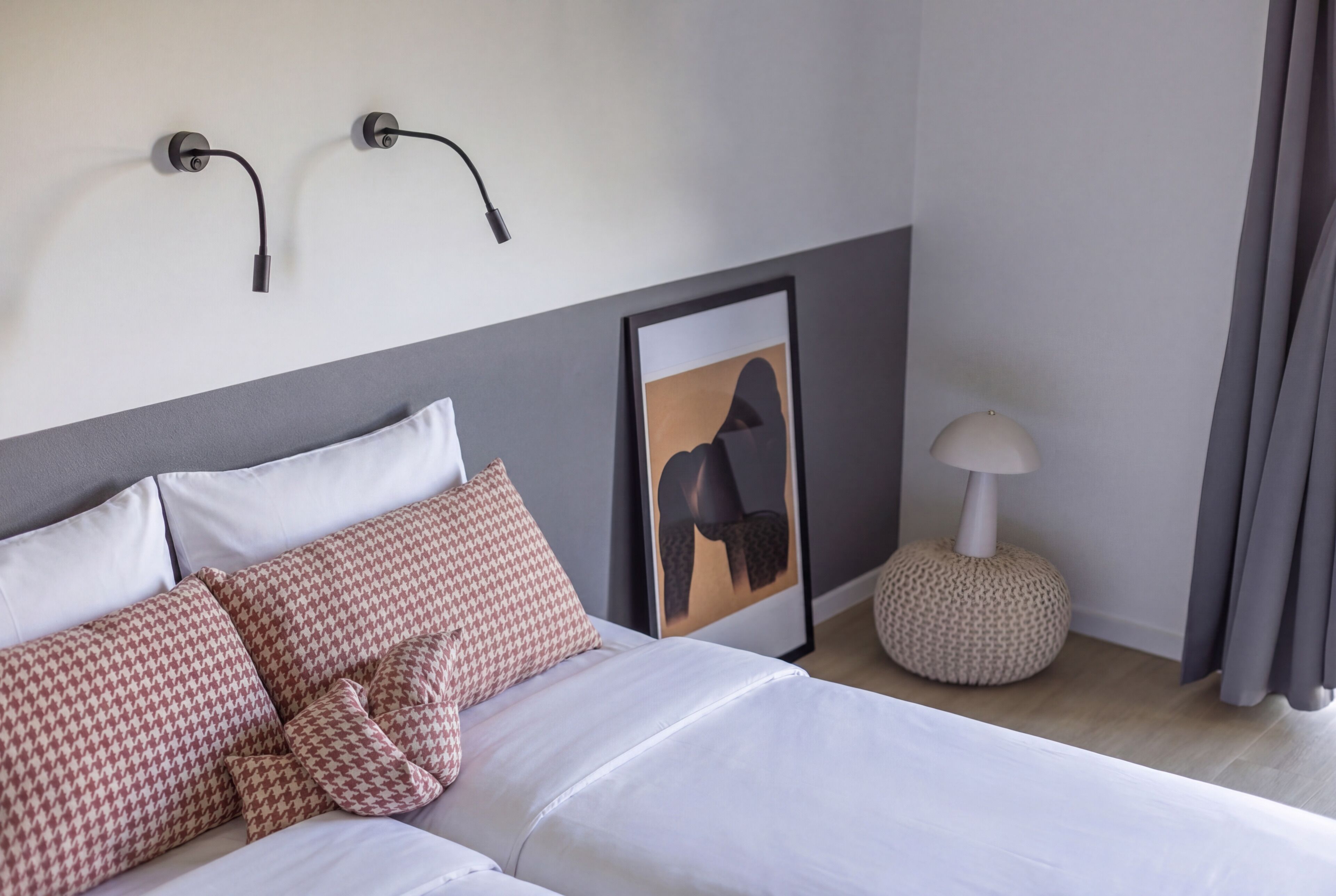 A neatly made bed with patterned pillows, a framed artwork on the wall, a floor lamp, and a round ottoman.