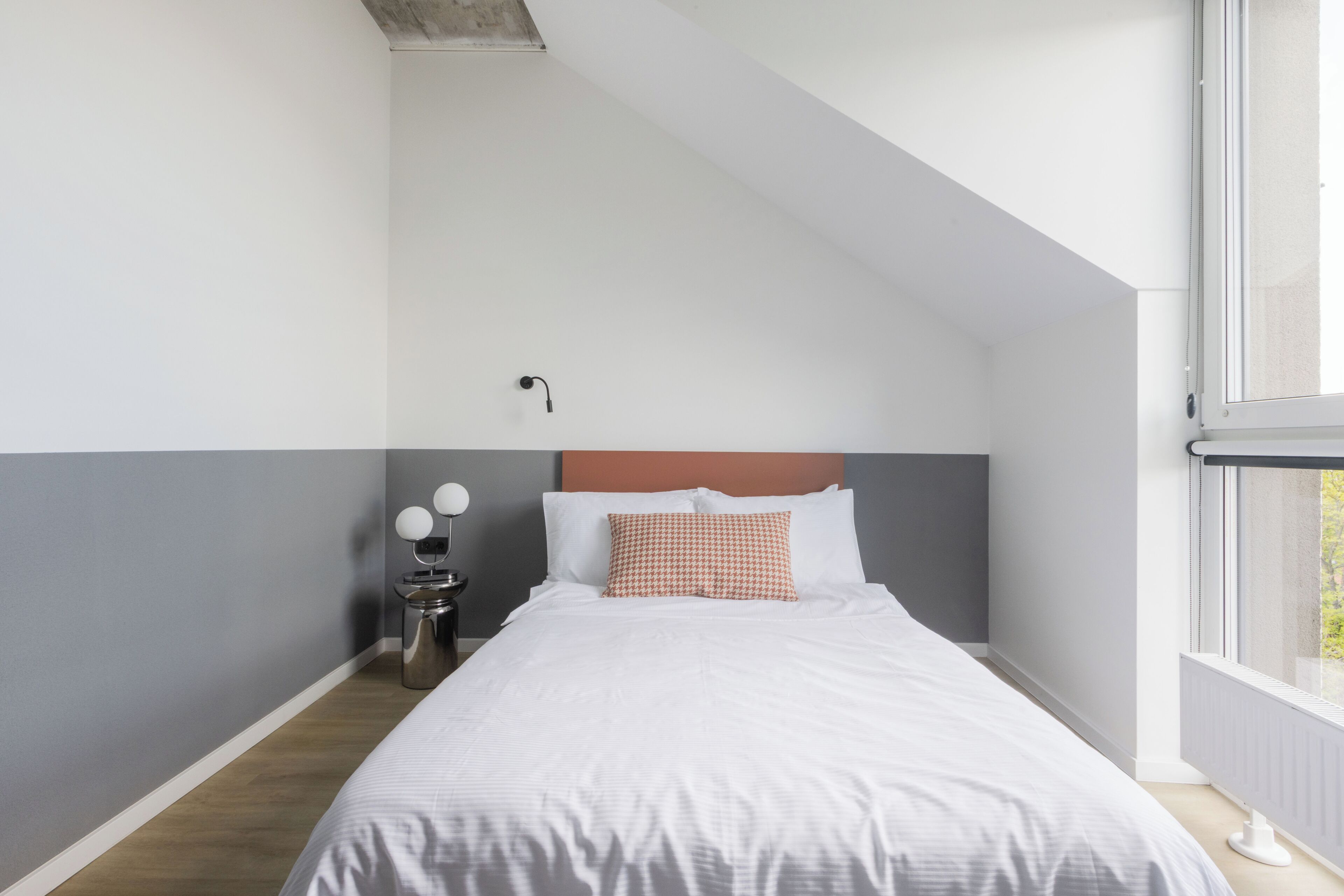 A bedroom with a bed, a nightstand with a lamp, a wall-mounted light, and a window.