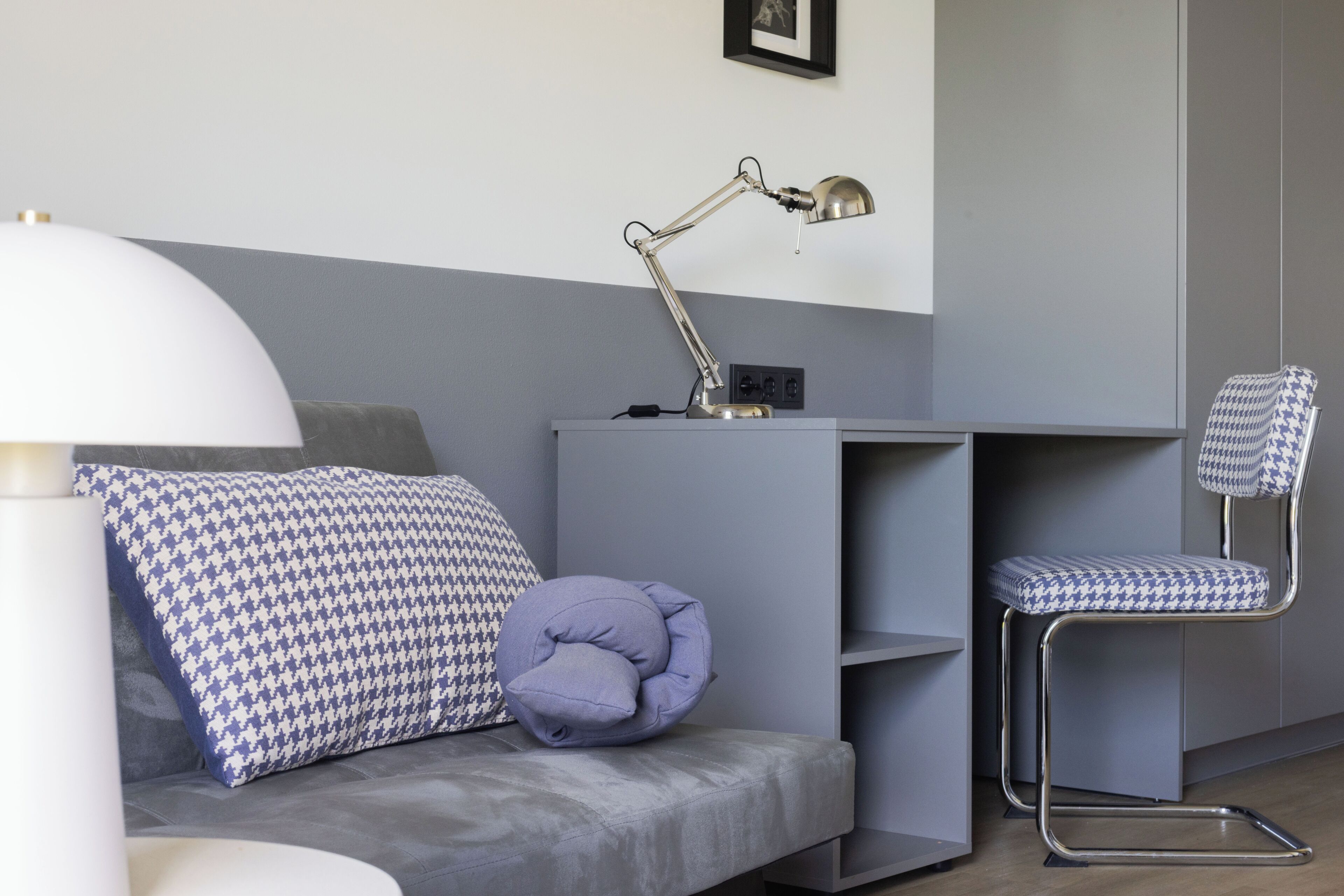 A modern room with a grey sofa, a patterned pillow, a desk with a lamp, and a houndstooth patterned chair.