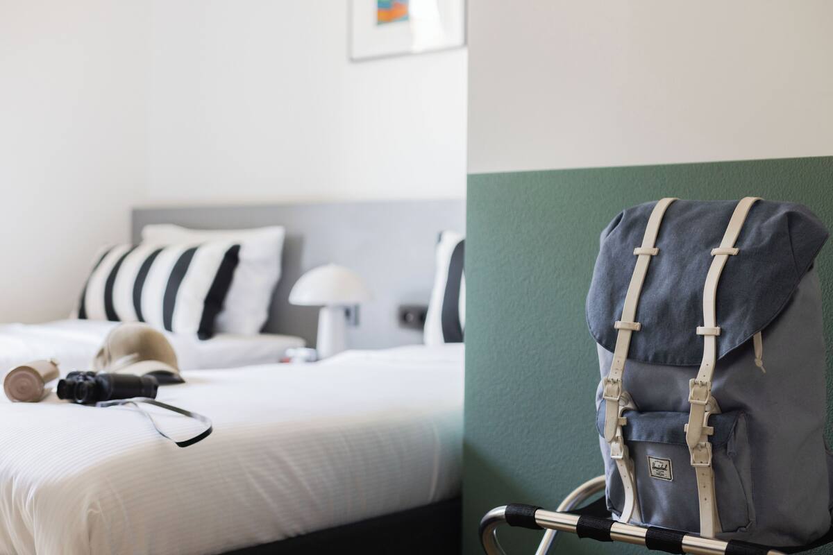 A hotel room with a bed, a bedside lamp, a suitcase, and a wheelchair.