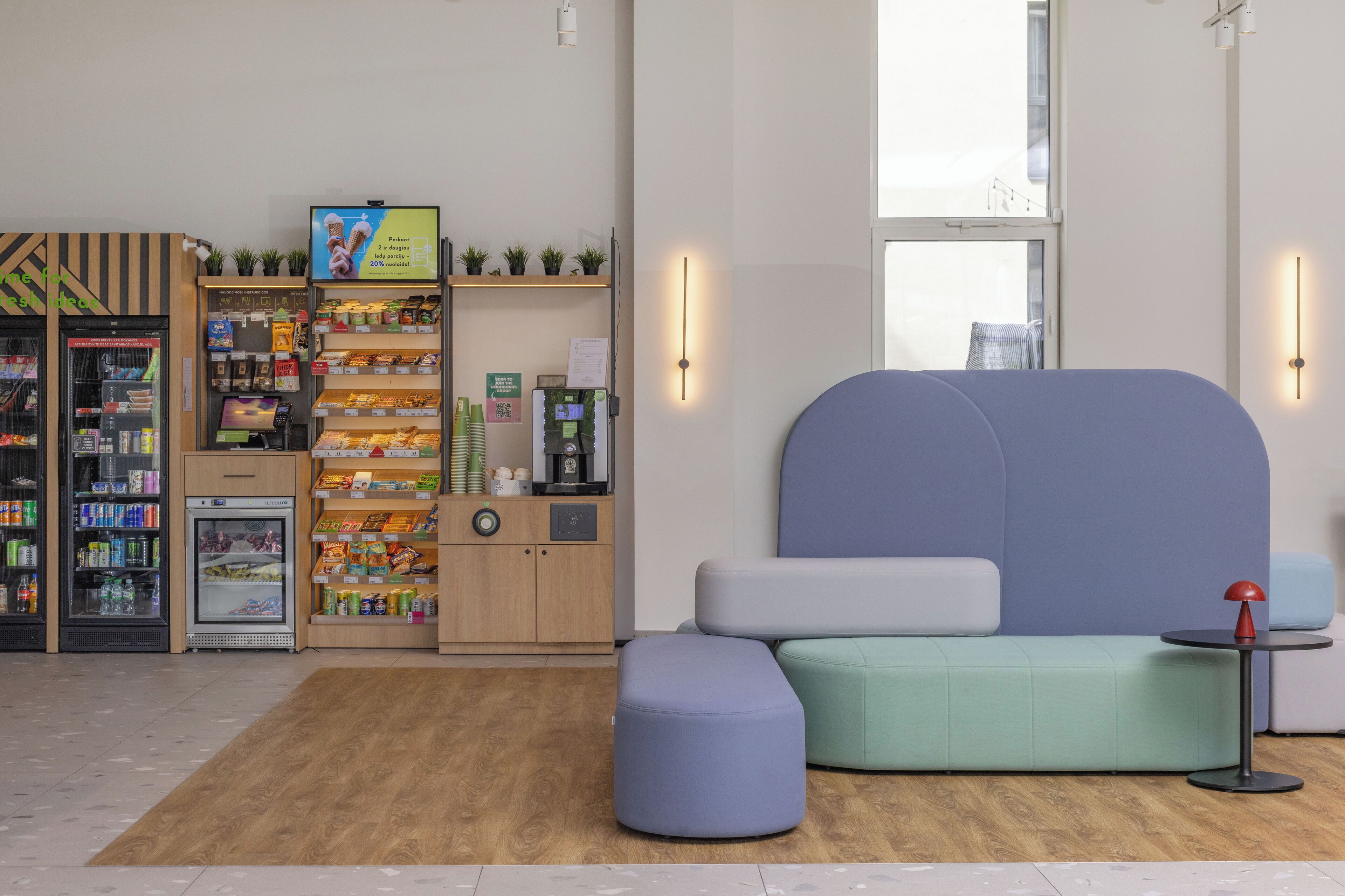 Un espacio comercial moderno y bien iluminado, con zona de estar, mostrador refrigerado y varias estanterías con productos.