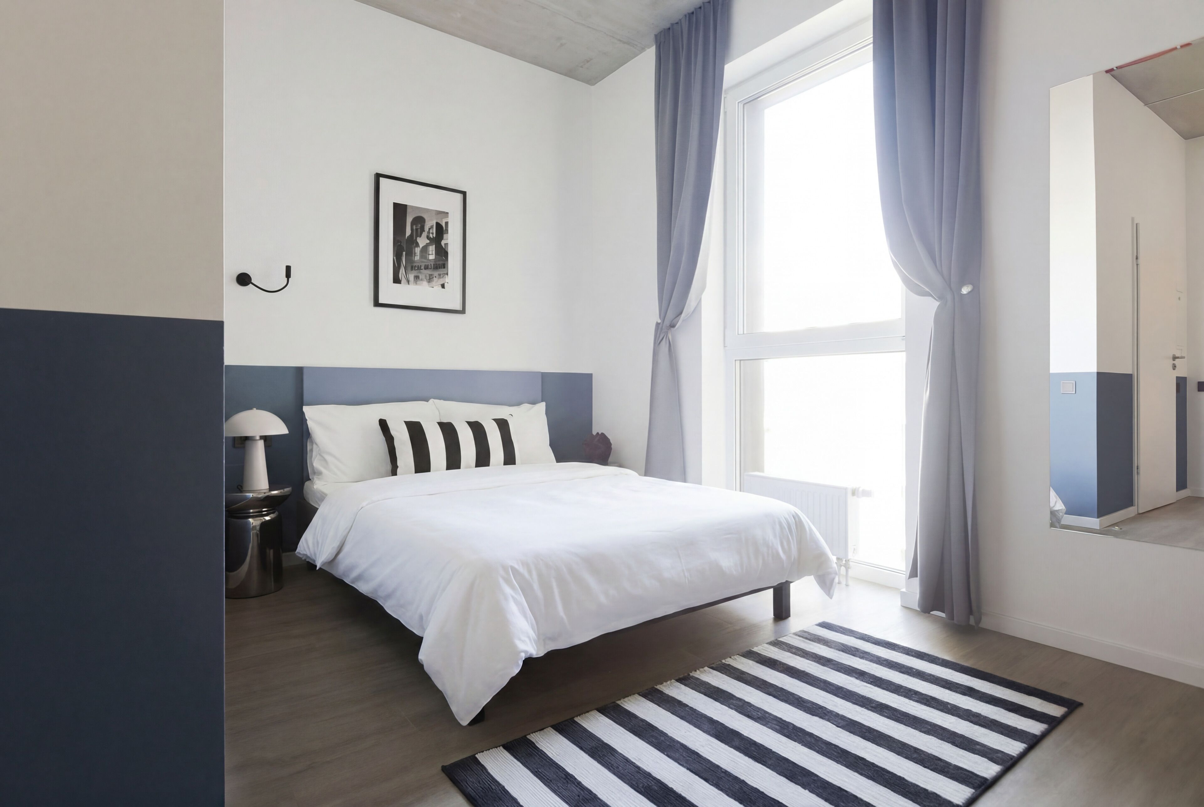 Een slaapkamer met een bed, nachtkastje, lamp, een lijst met een afbeelding en een raam met gordijnen.