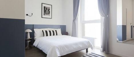 A bedroom with a bed, a bedside table, a lamp, a framed picture, and a window with curtains.