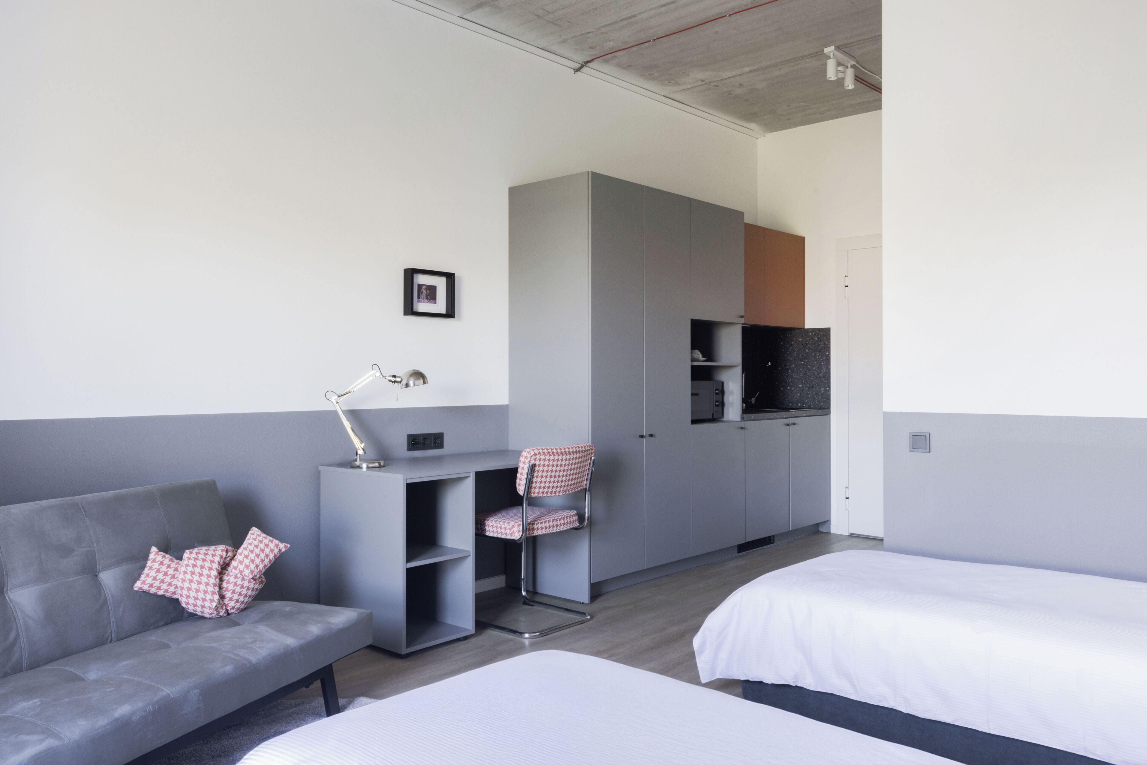 Une chambre d’hôtel moderne avec un canapé gris, deux lits simples, un bureau avec une chaise, une armoire et un coin cuisine.