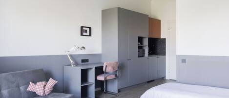 A modern hotel room with a grey sofa, two single beds, a desk with a chair, a wardrobe, and a kitchenette.