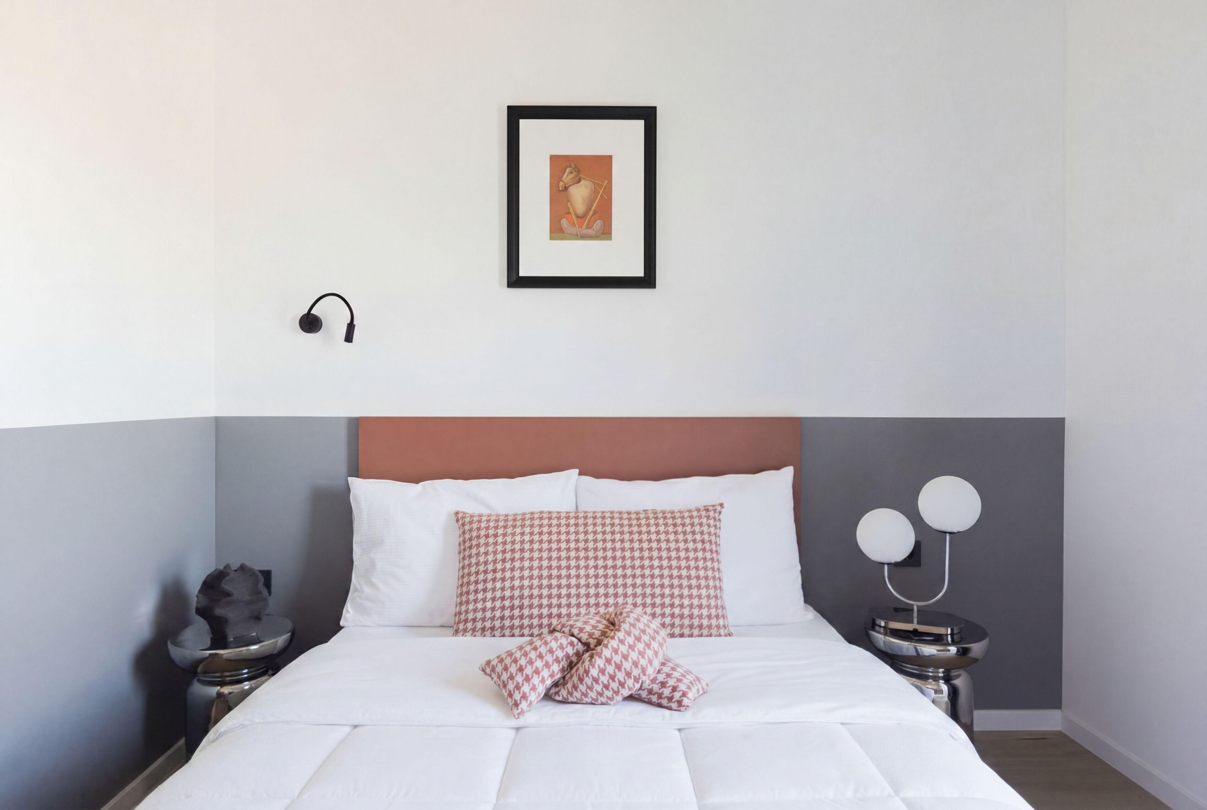 A neatly made bed with white bedding and a houndstooth pillow, a framed picture on the wall, and a bedside table with a lamp and a decorative item.