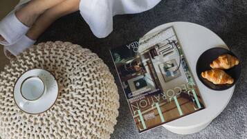 A person sitting on a grey couch, reading a magazine, with a knitted pouf and a plate of croissants on a small table beside them.