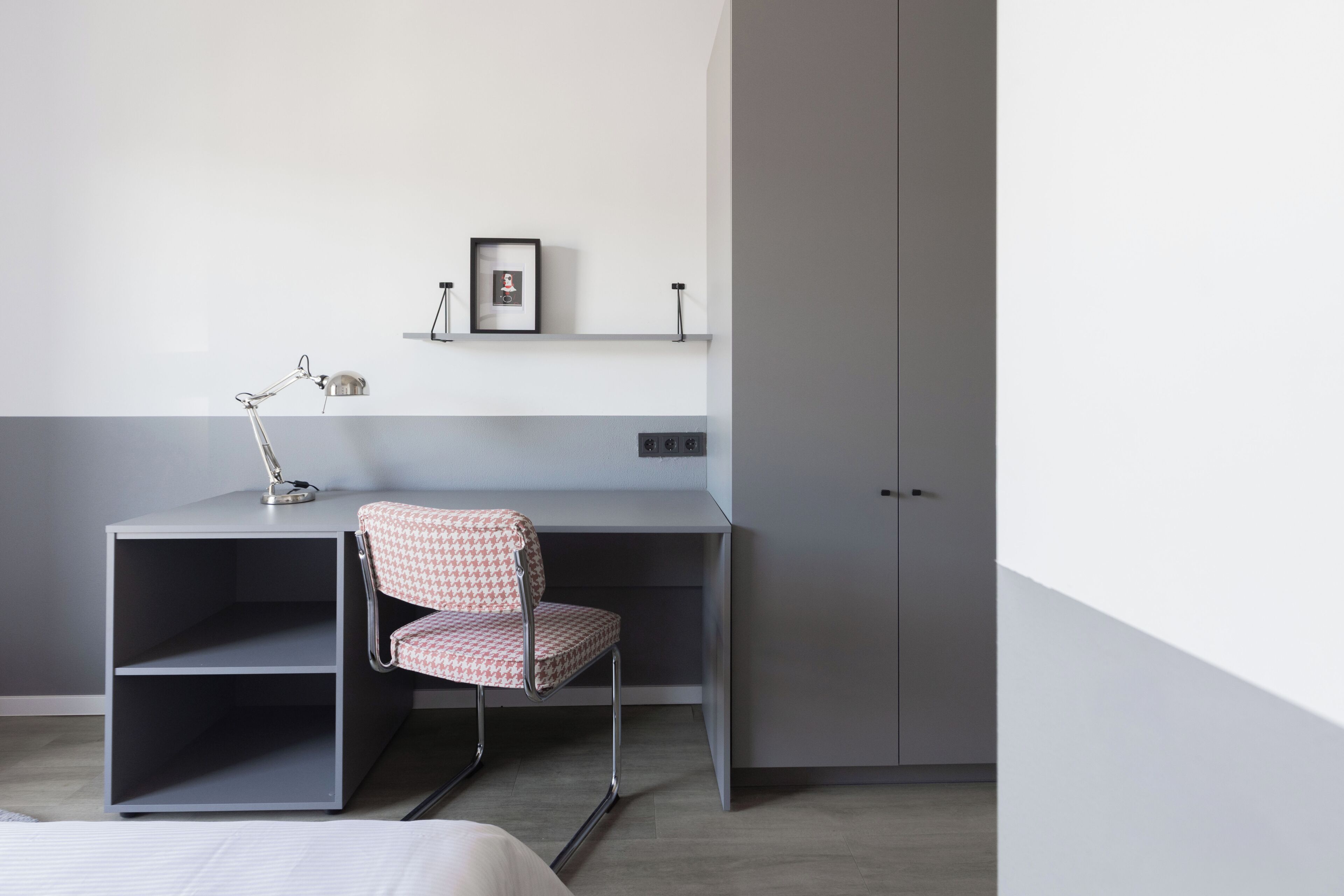 A modern room with a grey desk, a patterned chair, a lamp, and a wardrobe.