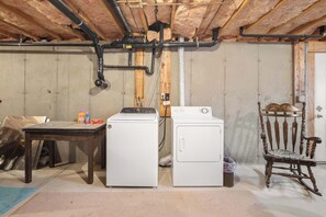 A laundry room with two white washing machines, a wooden table with cleaning supplies, and a rocking chair. - Game Room, Cozy & Mins to Beaches! (Eastham)