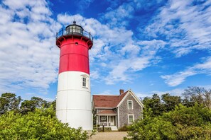 A lighthouse with a red and white tower, a small house with a porch, and lush greenery around. - Game Room, Cozy & Mins to Beaches! (Eastham)