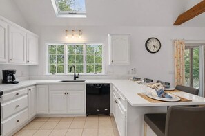 A modern kitchen with white cabinetry, a black dishwasher, a skylight, and a dining area with a table set for two. - Game Room, Cozy & Mins to Beaches! (Eastham)
