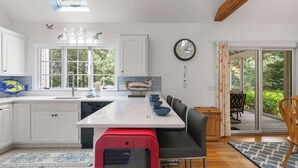 A modern kitchen with a central island, a dining area with chairs, and a view of the outdoors through large windows. - Game Room, Cozy & Mins to Beaches! (Eastham)