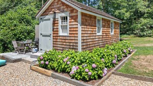 A small wooden shed with a door and windows, surrounded by a gravel area and flowering plants. - Walk to Water, Cozy, Dog Friendly! (Orleans)