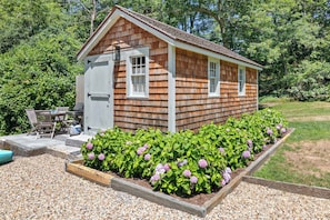 A small wooden shed with a door and windows, surrounded by a gravel area and flowering plants. - Walk to Water, Cozy, Dog Friendly! (Orleans)