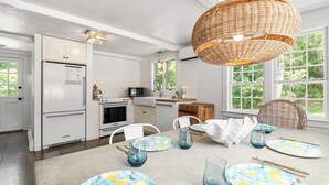 A modern kitchen with white cabinetry, a dining area with a table set for four, and large windows offering a view of greenery. - Walk to Water, Cozy, Dog Friendly! (Orleans)