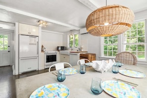 A modern kitchen with white cabinetry, a dining area with a table set for four, and large windows offering a view of greenery. - Walk to Water, Cozy, Dog Friendly! (Orleans)
