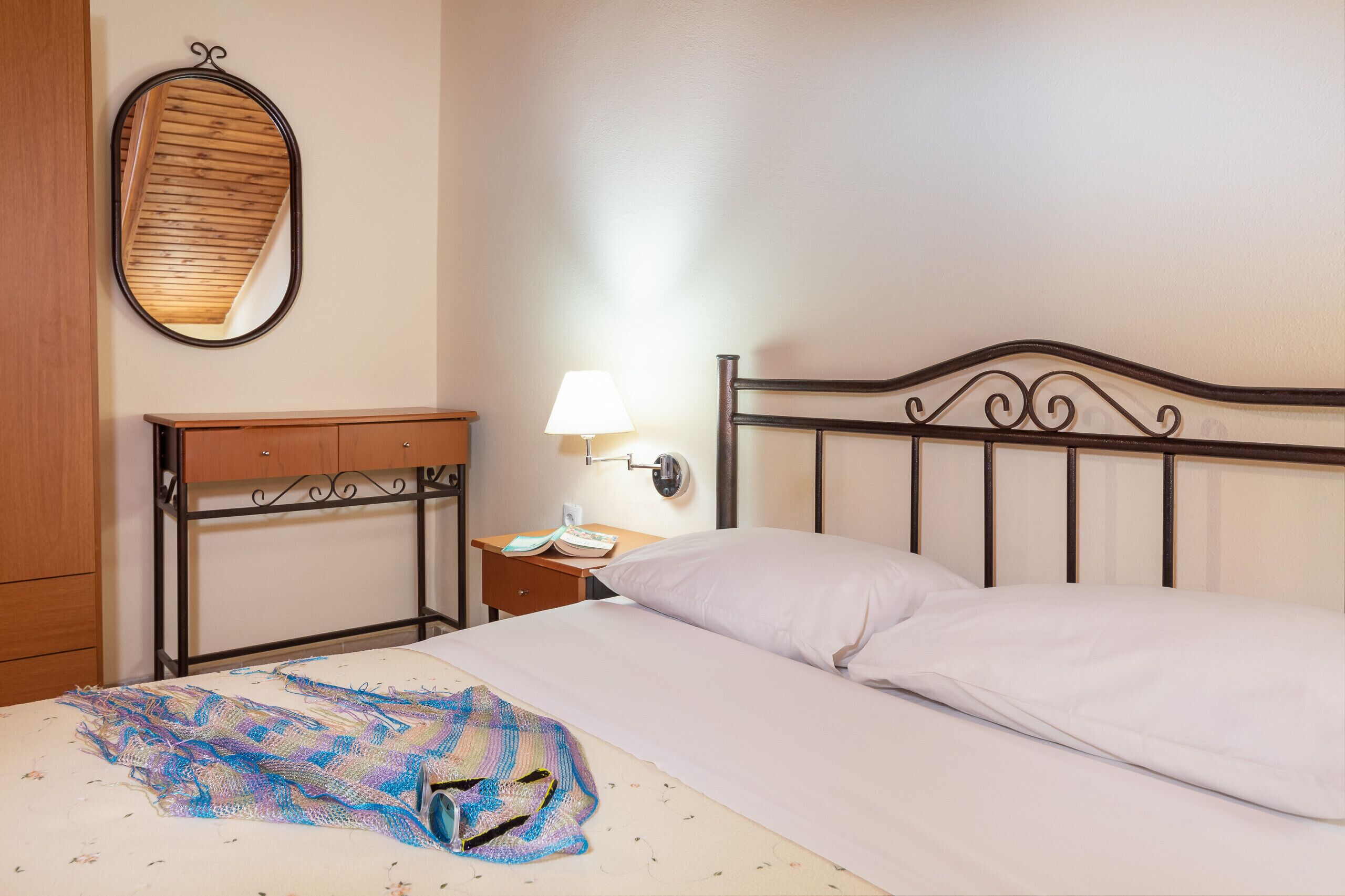 A bedroom with a bed, a nightstand, a lamp, a mirror, and a wooden wardrobe.