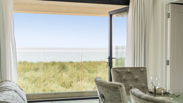Habitación con un amplio ventanal que da a una vista de la playa, con una silla acolchada y una mesita con un vaso.