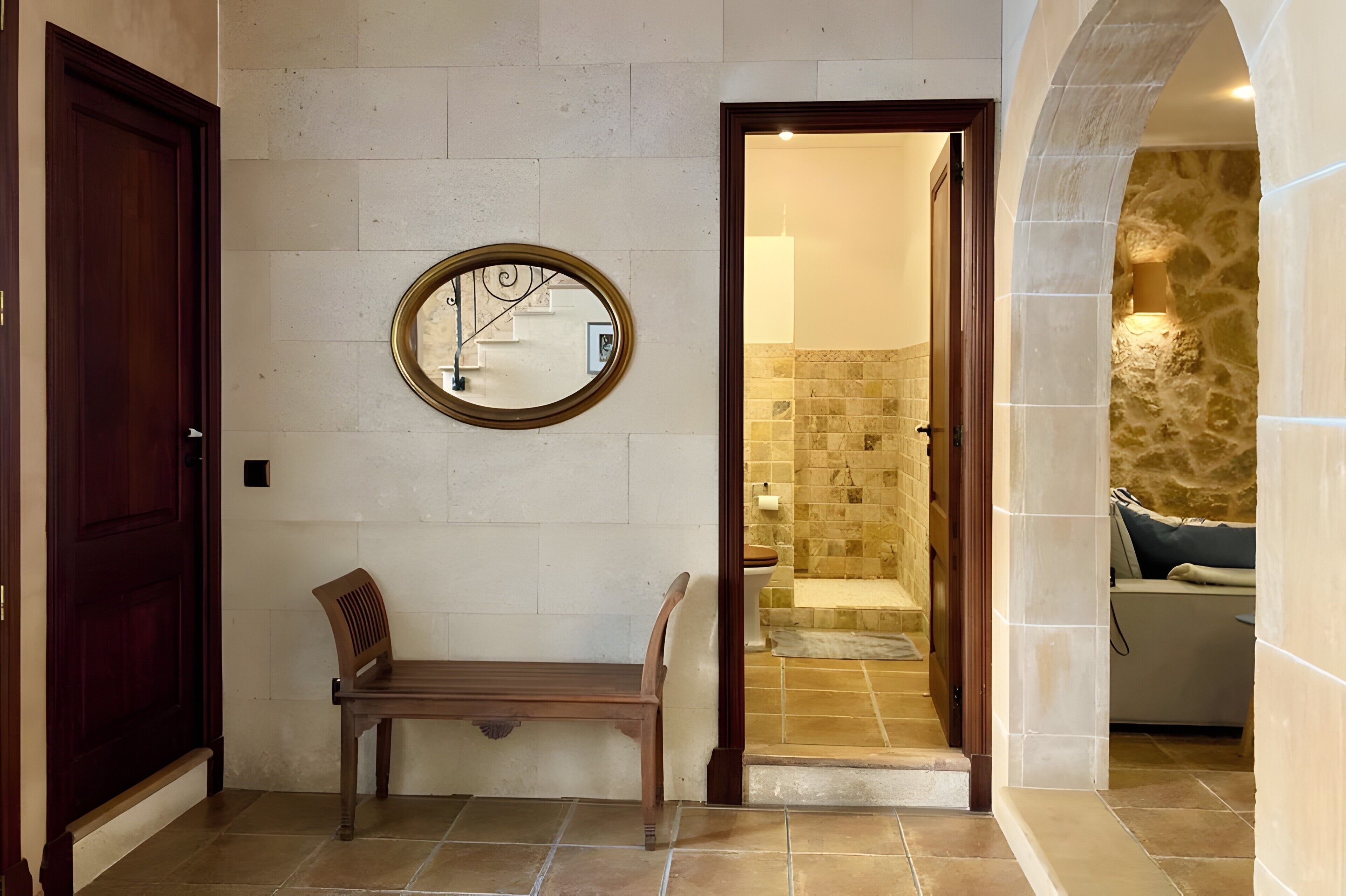 A hallway with a wooden bench, a round mirror, and a bathroom entrance with a tiled floor and a stone wall.
