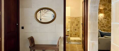 A hallway with a wooden bench, a round mirror, and a bathroom entrance with a tiled floor and a stone wall.