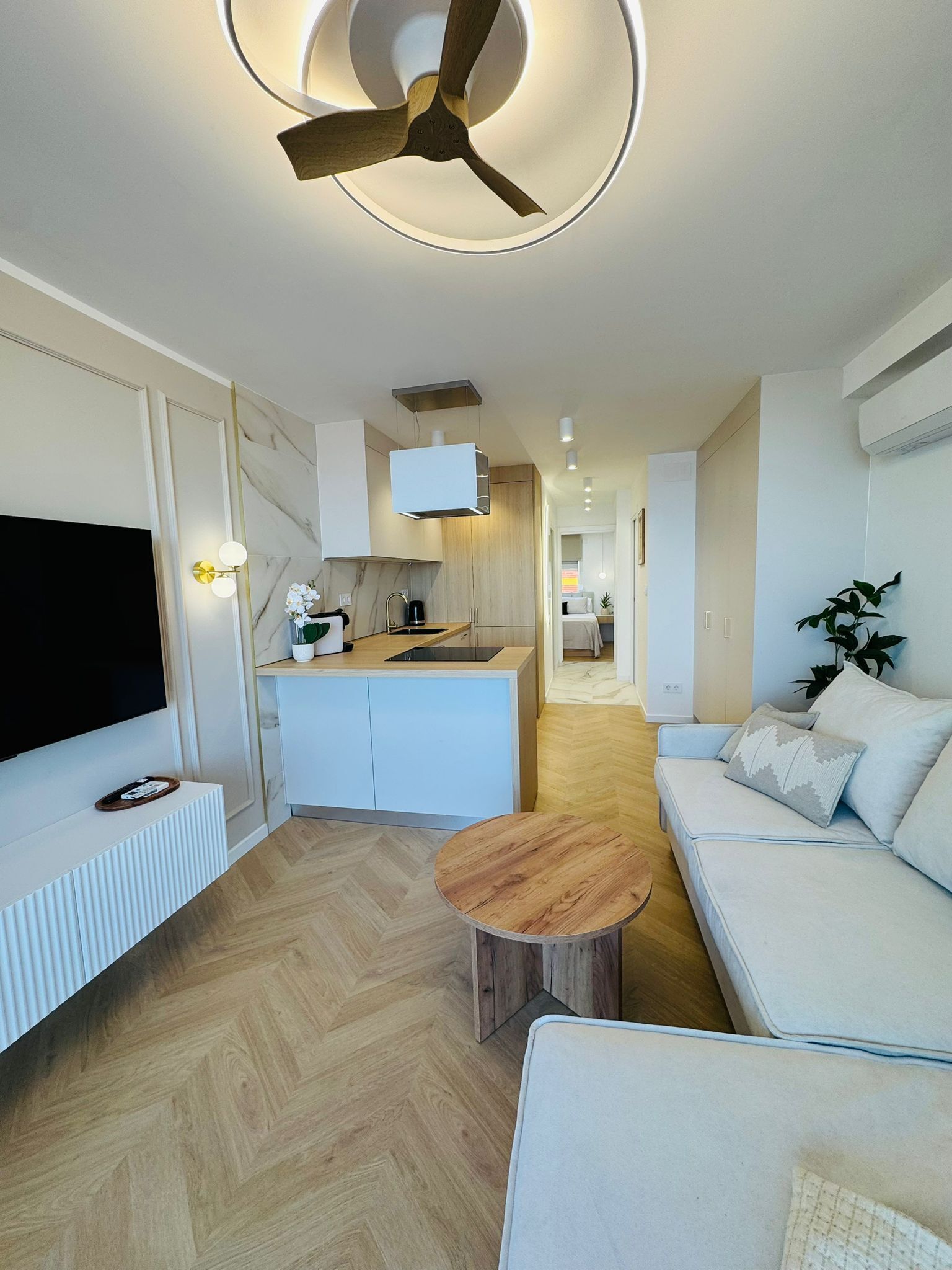 Una sala de estar moderna con un televisor de pantalla plana, una mesa de centro de madera, un sofá blanco y un ventilador de techo.
