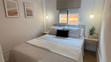 Un dormitorio con una cama doble, dos mesitas de noche, una ventana con persianas y dos lámparas de pared.