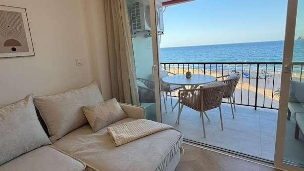 Ein modernes Wohnzimmer mit einer Couch, einem kleinen Holztisch und einem Balkon mit Blick auf Strand und Meer.