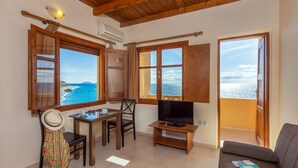 Una sala de estar acogedora con techo de madera, un televisor sobre un mueble bajo, un sofá y una mesa de comedor con sillas. Amplias ventanas ofrecen vistas al mar.