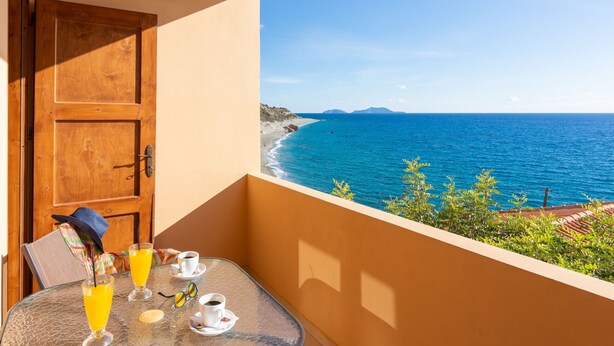 Un balcón con una mesa dispuesta para dos, con vistas a una playa de aguas azules cristalinas y montañas a lo lejos.
