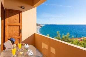 Ein Balkon mit einem Tisch für zwei Personen, Blick auf einen Strand mit klarem, blauem Wasser und entfernte Berge.