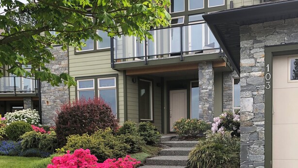 Ein modernes, zweistöckiges Haus mit Naturstein-Fassade, einem Balkon und einem gepflegten Garten mit rosa Blumen.