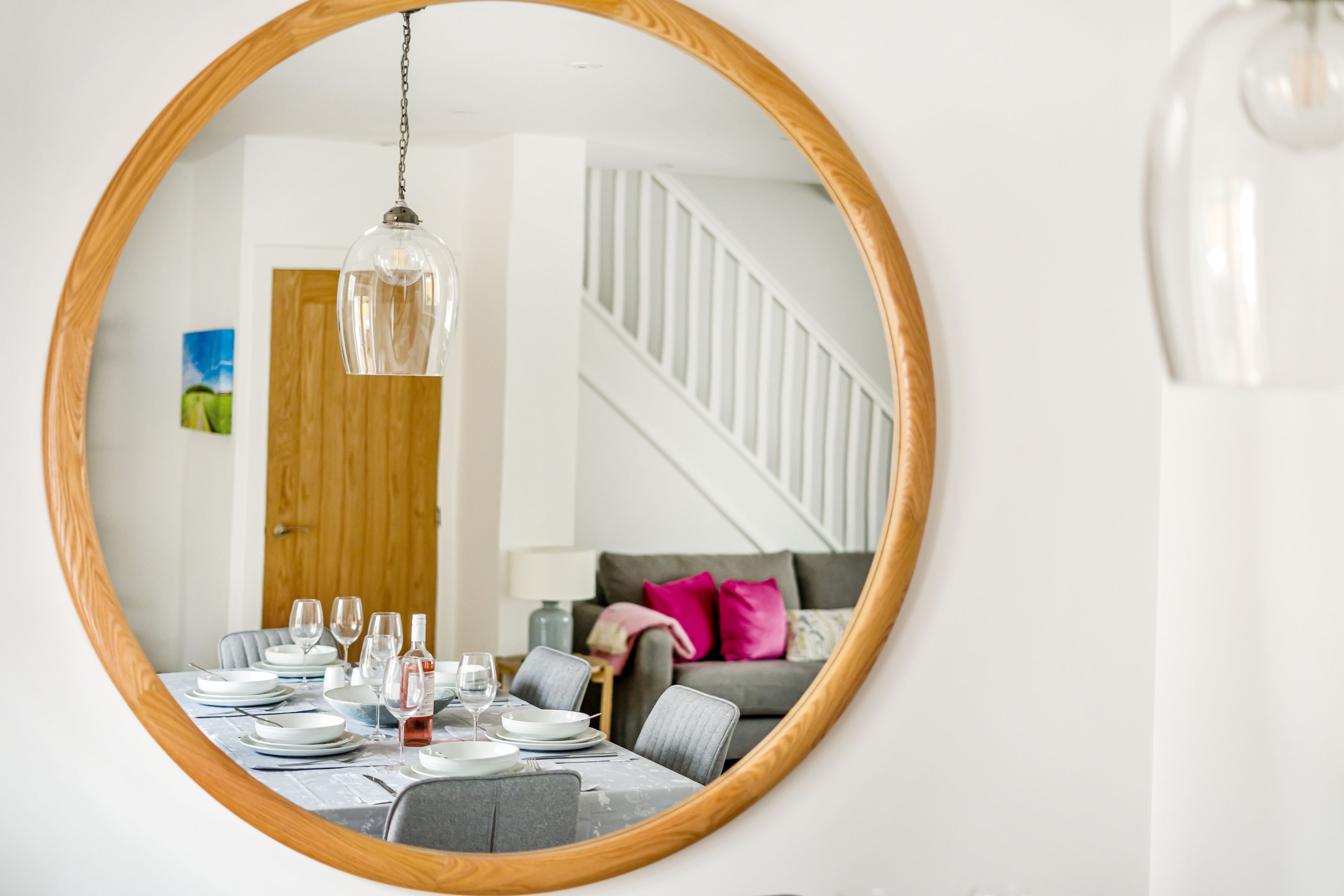 A dining area with a round mirror reflecting a staircase, a dining table set with plates, glasses, and a bottle, and a grey sofa with pink pillows in the background.